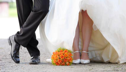 Männerbeine im schwarzen Anzug. Frauenbeine: Unterschenkel sichtbar, der Rest vom weißen Hochzeitskleid verdeckt. Orangene Blumen liegen zwischen dem Brautpaar auf dem Boden.