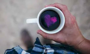 Ein Mann hält eine weiße Tasse vor seinem Bauch. Die Sicht von oben läßt schwarzen Kaffe mit einem lilafarbenen Herz in der Mitte erkennen.
