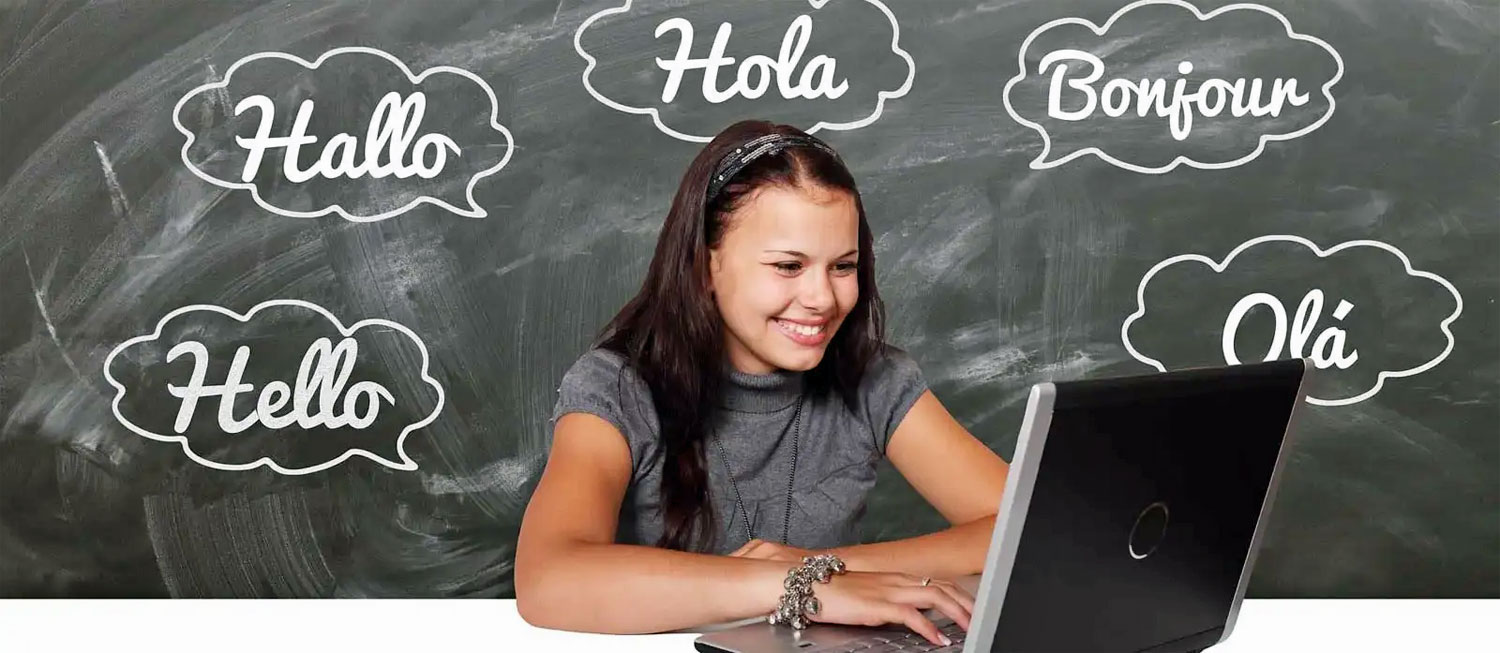 Junge, lächelnde Frau schaut auf einen geöffneten Laptop. Im Hintergrund eine Tafel mit den Aufschriften: Hello, Hallo, Hola, Bonjour, Olá.