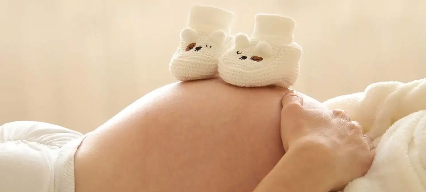 Bauch einer auf den Rücken liegenden hochschwangeren Frau. Auf dem Bauch ein Paar süßer Babypantoffeln in weiß.