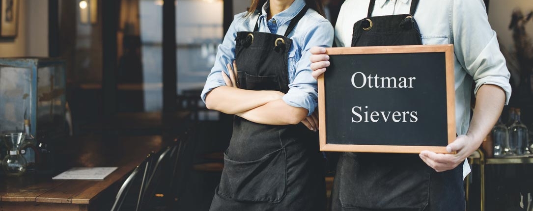 Frau und Mann mit schwarzer Schürze halten in einem Restaurant eine Tafel in der Hand. Darauf steht: Ottmar Sievers.